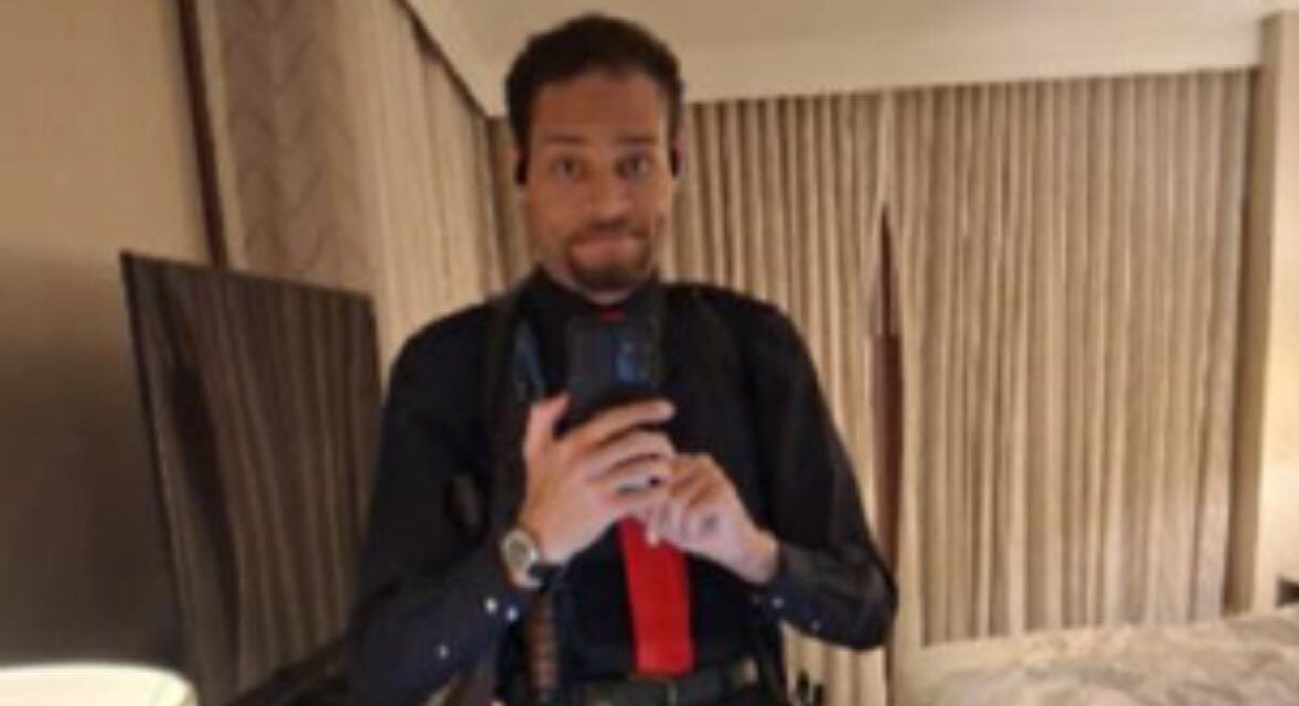 Με μαύρο πουκάμισο, κόκκινη γραβάτα και ζωσμένος με όπλα ποζάρει χαμογελώντας πριν πυροβολήσει